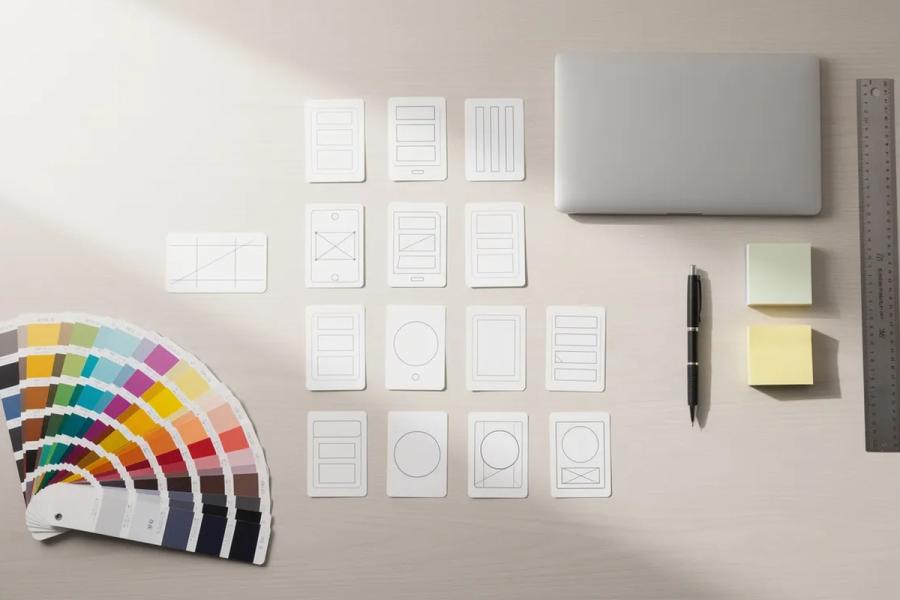 Colour swatches and wireframe layout cards arranged on a desk beside a laptop, pen, sticky notes and ruler